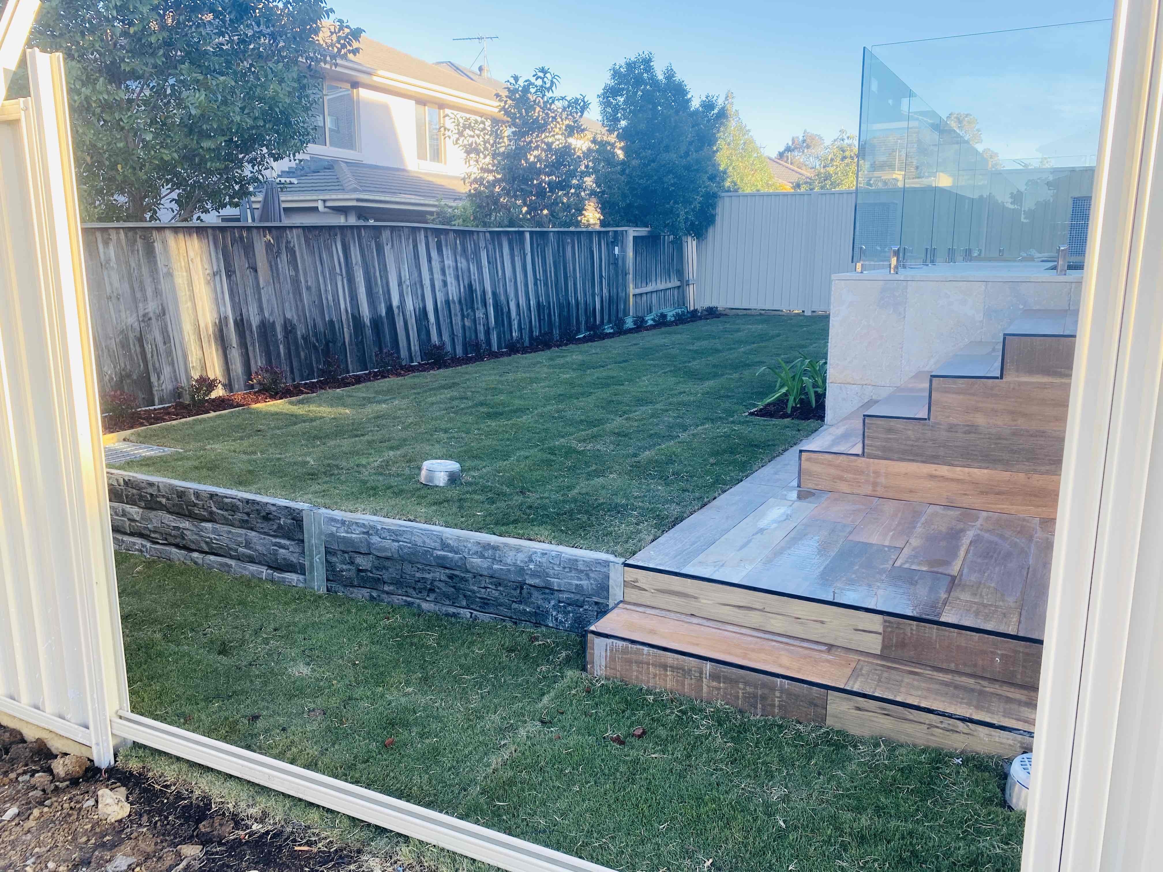 Finished backyard with new turf, a grey block retaining wall and tiled steps up to a glass-fenced pool