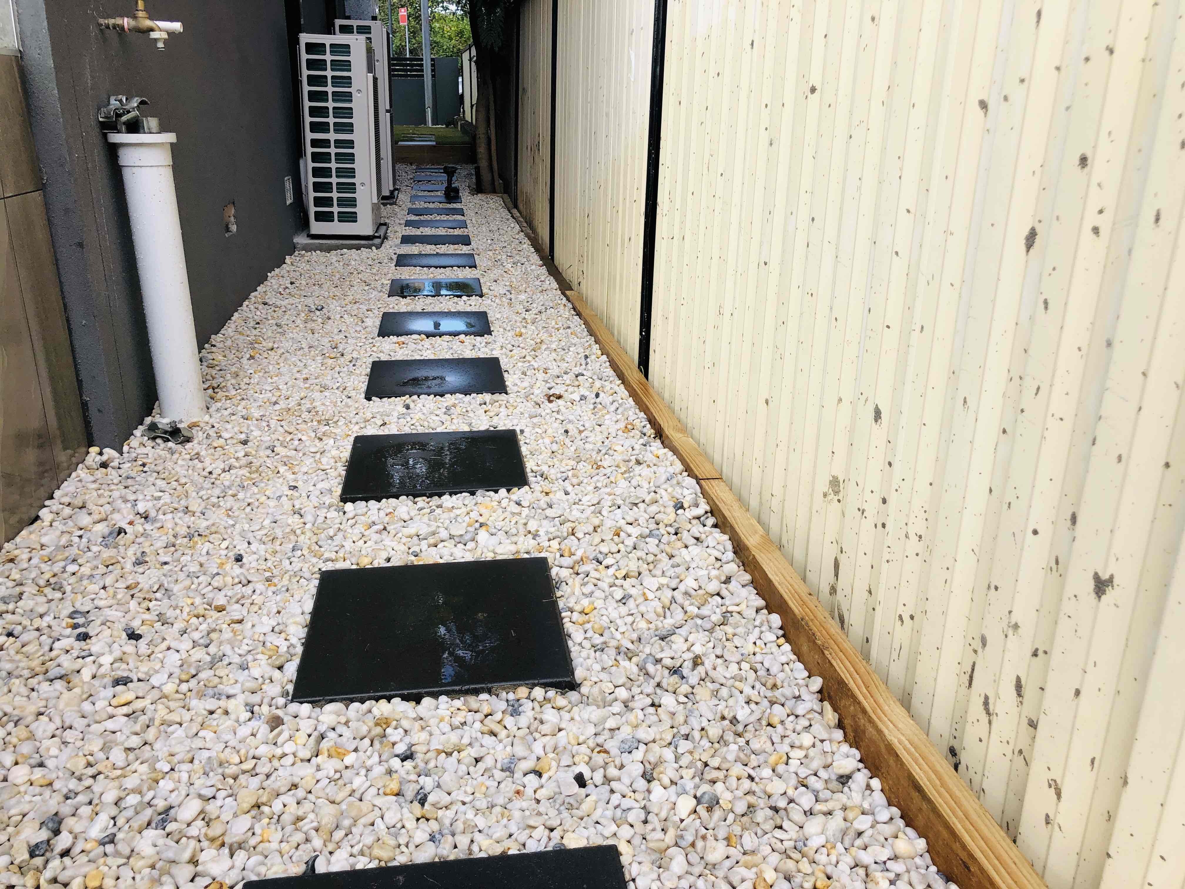 Black square pavers set into white pebbles down a side passage beside a cream colorbond fence